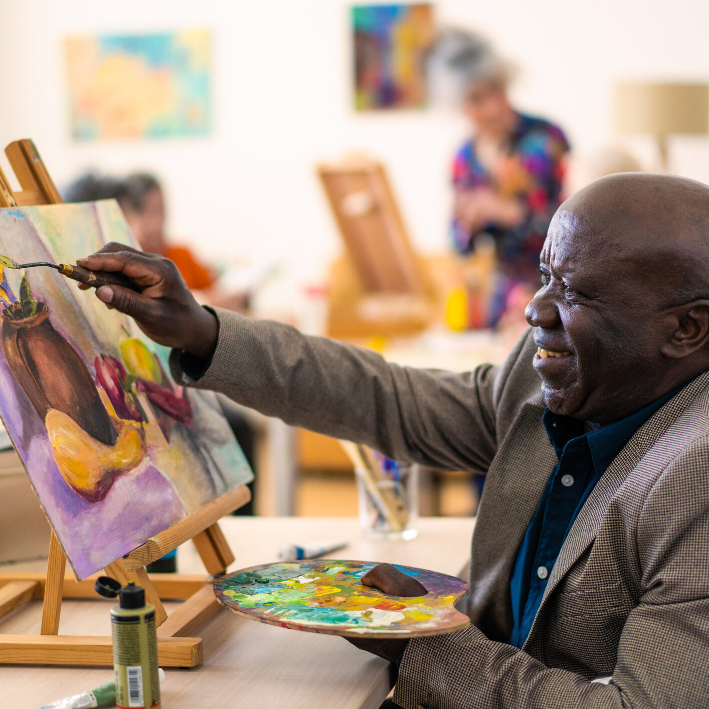 A senior African-American man is painting a picture using a paintbrush and palette for his composition on the canvas, behind him other members of the community are also painting
